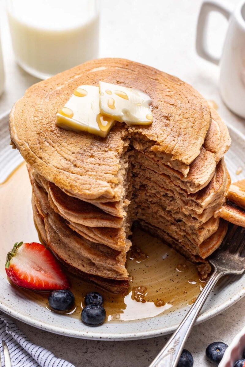 A stack of oat flour pancakes topped with pats of butter and syrup with fruit on the side and a large slice taken out of the stack.